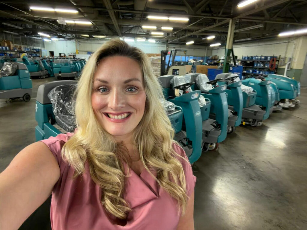 PowerKleen team member standing inside a commercial cleaning equipment warehouse with multiple floor scrubber machines in the background.