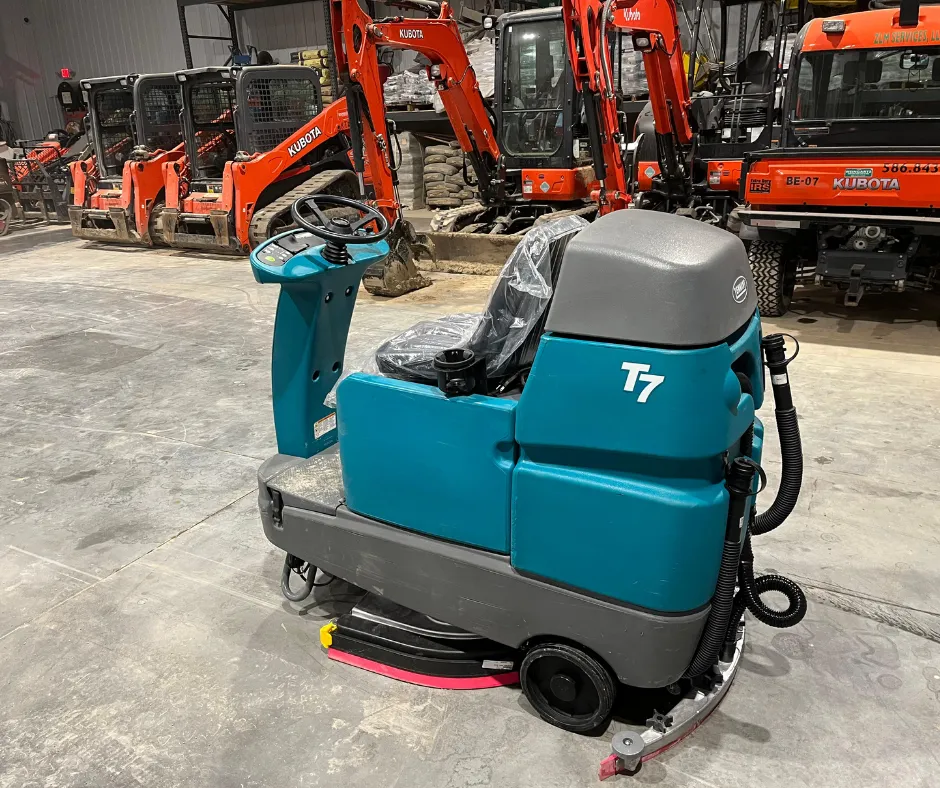 PowerKleen Tennant T7 ride-on floor scrubber inside a heavy equipment facility with Kubota construction machinery and industrial equipment in the background.