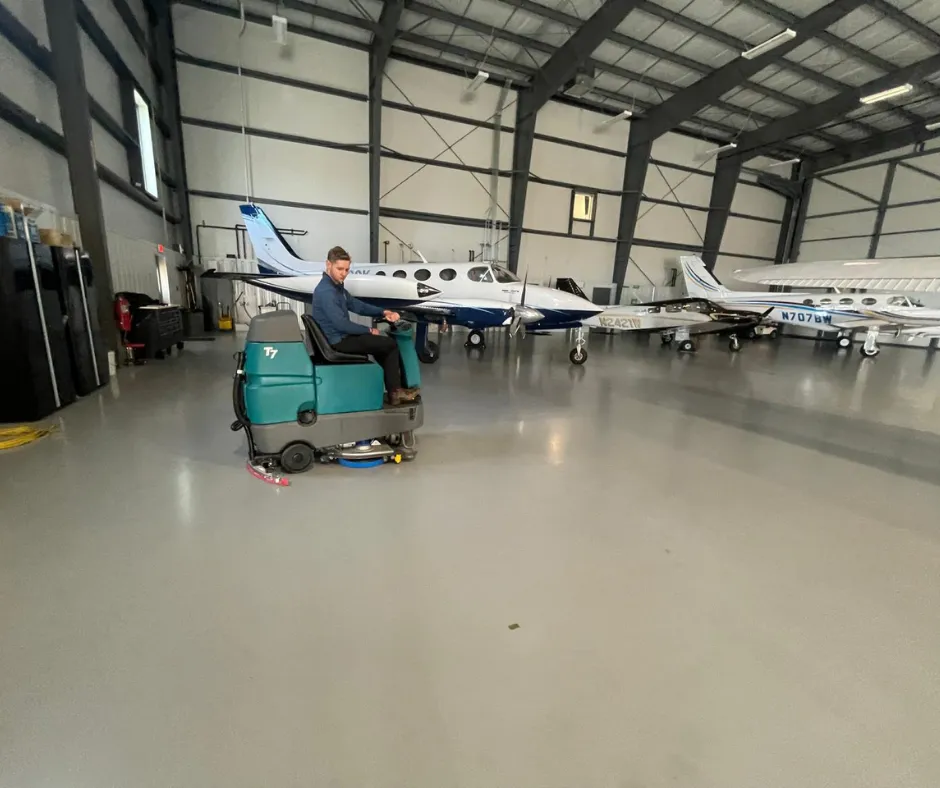 PowerKleen Tennant T7 ride-on floor scrubber cleaning polished floors inside a private aircraft hangar with airplanes in the background.