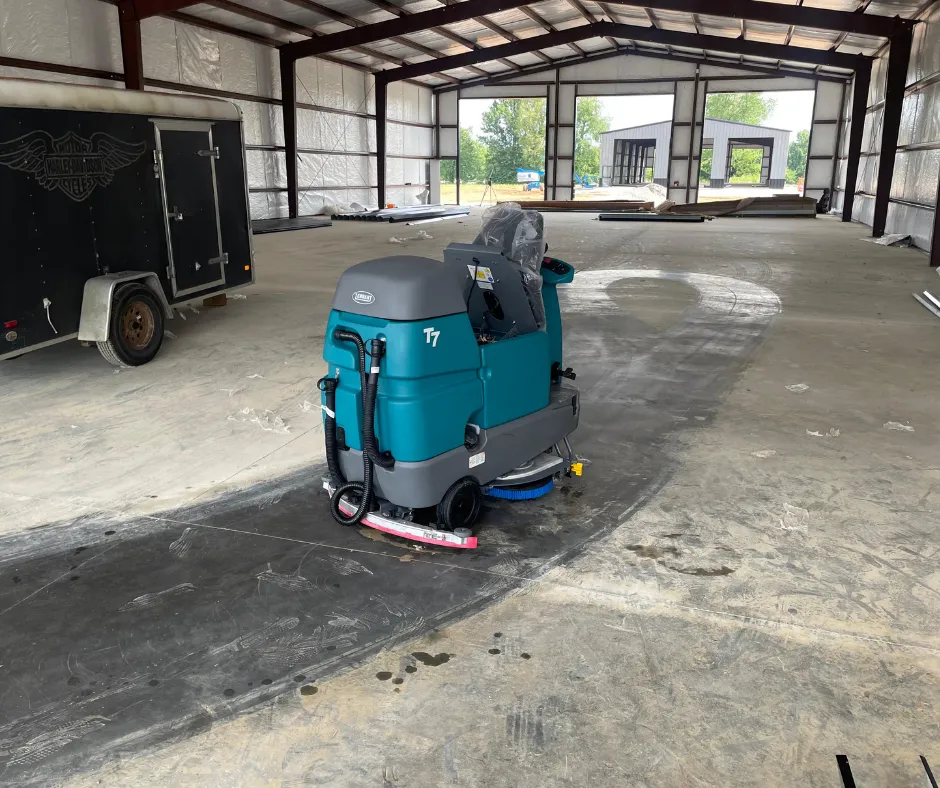 PowerKleen Tennant T7 ride-on floor scrubber cleaning concrete floors inside a large industrial warehouse and commercial workspace.