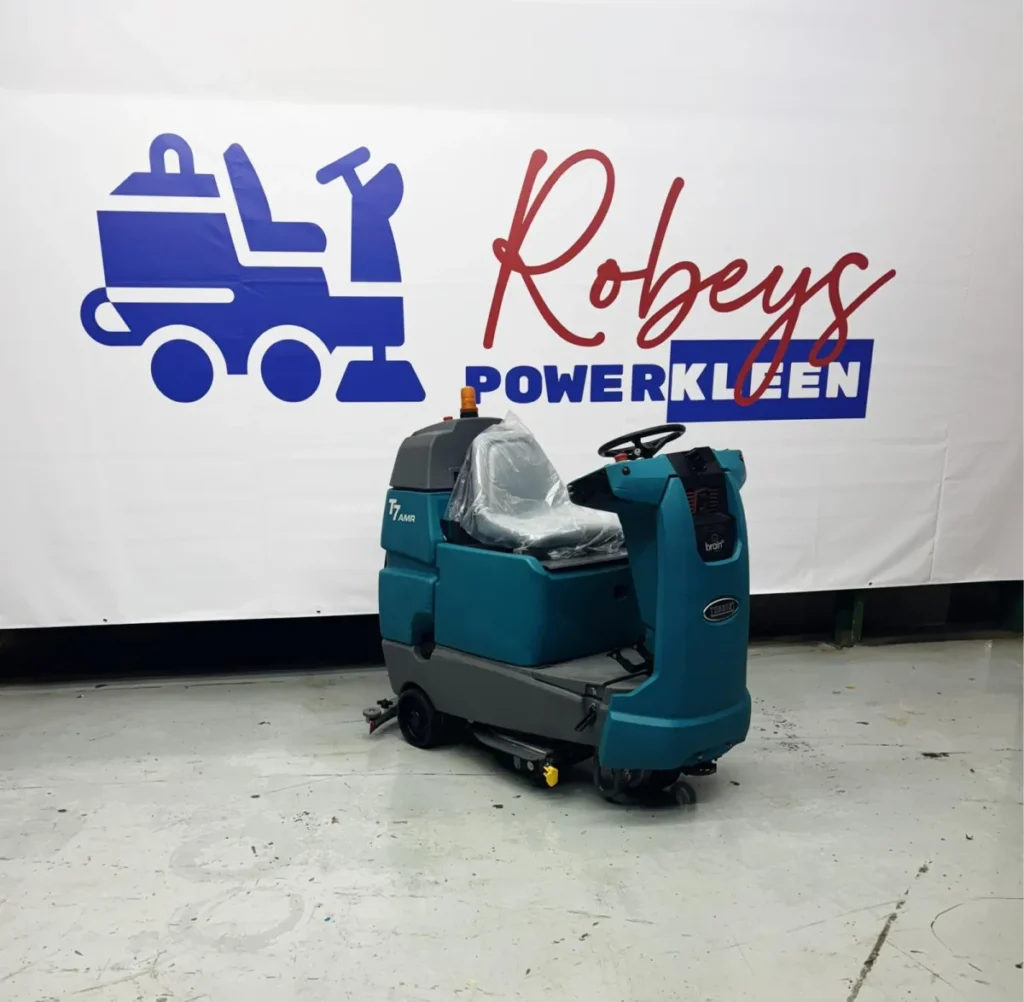 Front-right angled view of a Tennant T7 AMR autonomous rider floor scrubber displayed indoors in front of a Robey’s PowerKleen banner, highlighting its compact industrial cleaning design and automated floor scrubbing system.